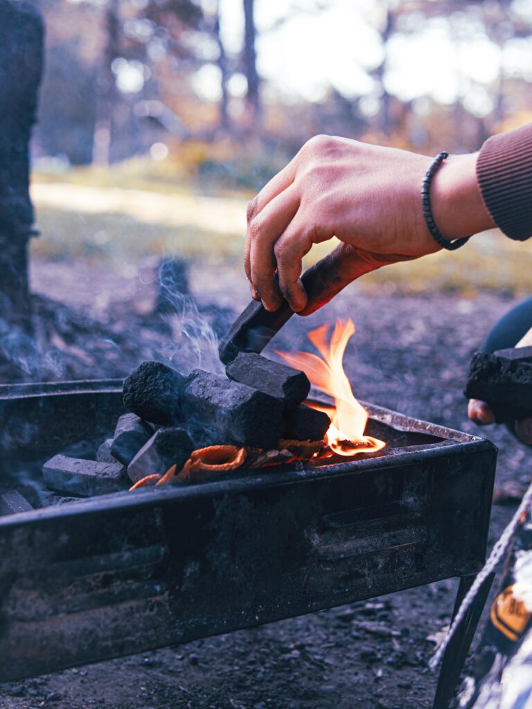 Hands wearing bracelet igniting charcoal on an outdoor grill, creating a warm fire.